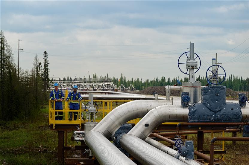 Two workers stand on a platform at Suncor's Firebag site, overlooking pipelines, valves and processing equipment. Additional piping infrastructure, boreal forest and a cloudy blue sky are visible in the background.