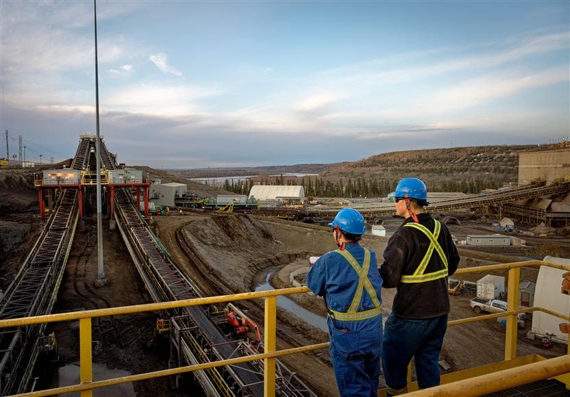 A female and a male worker standing at a railing on the top of Plant 82 Extraction facilities looking out towards the river. They are wearing blue hard hats and safety glasses. 