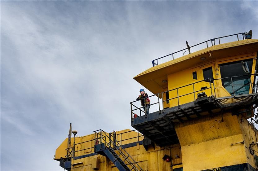 Un travailleur de Suncor se tient debout et appuyé sur le garde-corps d’une plateforme devant la cabine d’opérateur d’une pelle électrique à l’usine de base. Il porte un casque bleu, des lunettes de sécurité, une veste haute visibilité et des gants.