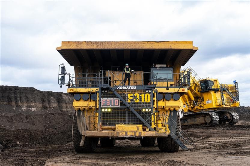 A haul truck operator stands on the front deck of a Komatsu 980E haul truck, of which Suncor has the largest fleet in the world - in the mine operations at Suncor's Fort Hills site. The operator is wearing full PPE and smiling towards the camera. In the background is another haul truck and cloudy grey skies.