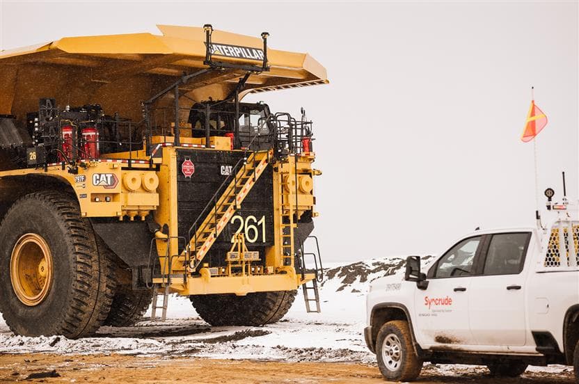 Un camion de transport autonome à la mine Mildred Lake de Syncrude. Un véhicule de parc blanc décoré avec un logo de Syncrude est stationné à côté.