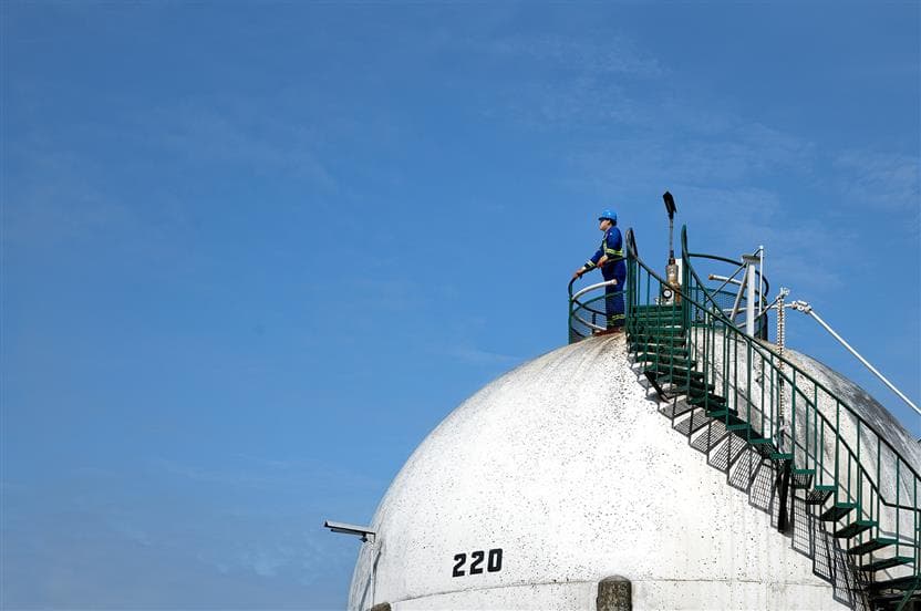 Un travailleur portant un casque de sécurité bleu, des lunettes de sécurité, une combinaison et des gants debout sur le dessus d’une sphère de butane au terminal de Burrard, situé sur la côte ouest de la Colombie-Britannique.