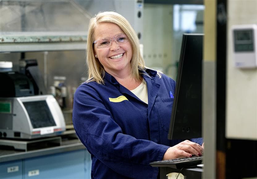 Photo portrait d’une technicienne de laboratoire travaillant à un ordinateur dans le laboratoire de l’usine de base. Elle porte des lunettes de sécurité et une blouse de laboratoire et sourit à la caméra.