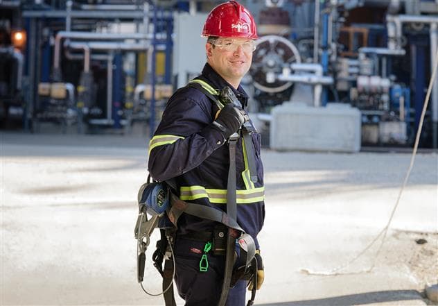 A male worker standing outside at the Edmonton refinery. He is smiling towards the camera, and is wearing a red hard hat, safety glasses, gloves, and a high-visibility jacket.