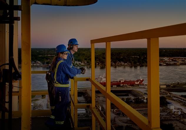 Two workers stand at a railing on top of a tower looking out at the Sarnia Refinery. They are wearing blue hard hats, coveralls, safety glasses and gloves. It is sunrise and the St. Clair River is in the background.