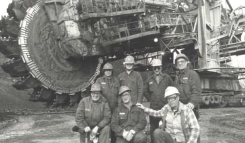 A group of seven Sun Oil employees stand in front of one of the giant bucketwheel excavators used for mining at the time. The employees all wear hard hats.