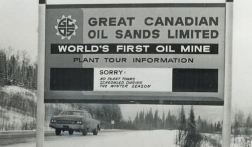 A sign that reads “Great Canadian Oil Sands Limited, World’s First Oil Mine”. It also offers some information on tours of the plant. A car is passing in the background.