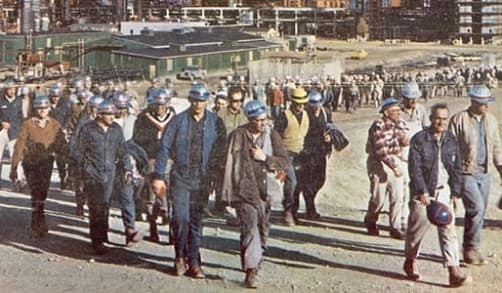 A large group of employees arrive for work at a Suncor site in the morning. Many are wearing blue hard hats and work clothes.