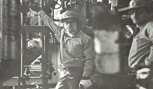 Two employees stand in the plant in northern Alberta. Both wear hard hats, button-up shirts and gloves.