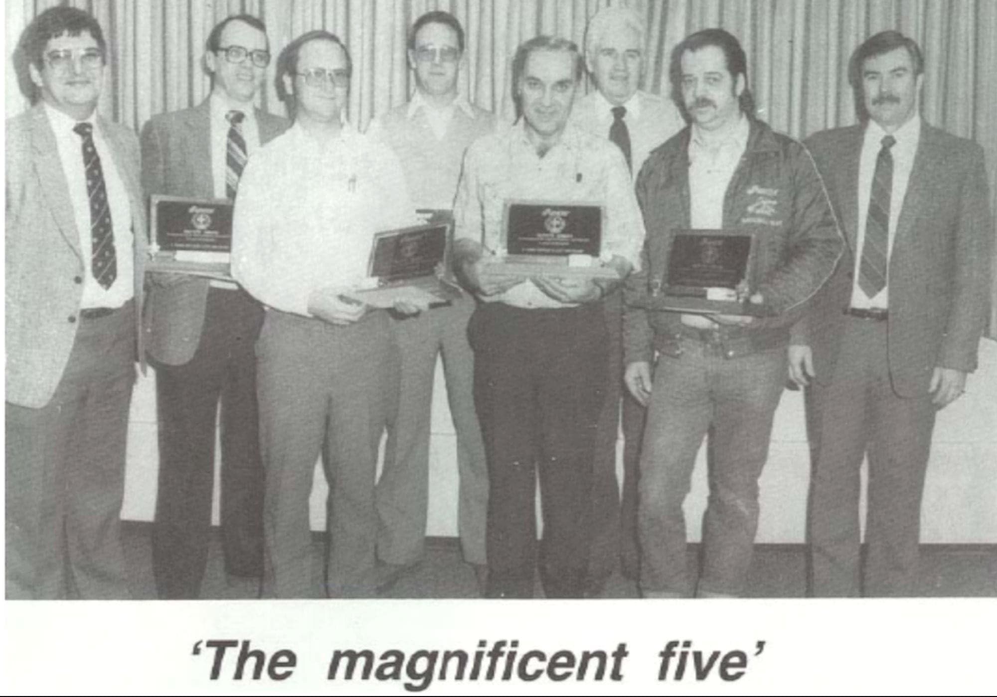 A group of eight men in suits pose for a photo. Four men hold awards and a caption reads ‘The magnificent five’.