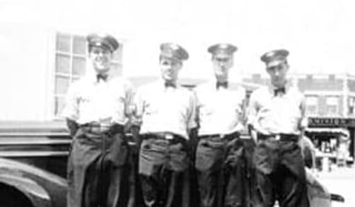 Four male Sun Oil service station attendants are pictured in front of a car. They are wearing hats, white shirts, bow ties and jeans.