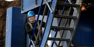 Un travailleur se tient dans les escaliers d’embarquement d’une pelle électrique dans la mine. Son bras repose sur la rampe et il porte un casque de protection bleu, des lunettes de sécurité et des gants.
