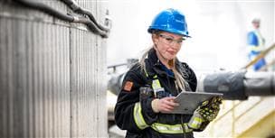 A worker standing outside at Plant 800 at MacKay River. She is wearing a blue hard hat, coveralls, safety glasses and gloves. In her hands she's holding a Microsoft Surface Pro. There is another worker (blurred out) standing in the background.