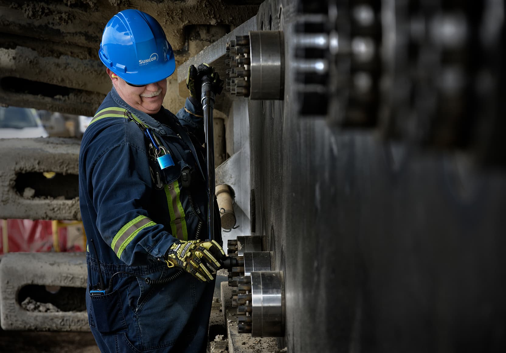 A male worker is working on an electric shovel track on the Borealis Shovel Pad at Base Plant. He is wearing a blue hard hat, ear plugs, coveralls and safety glasses.