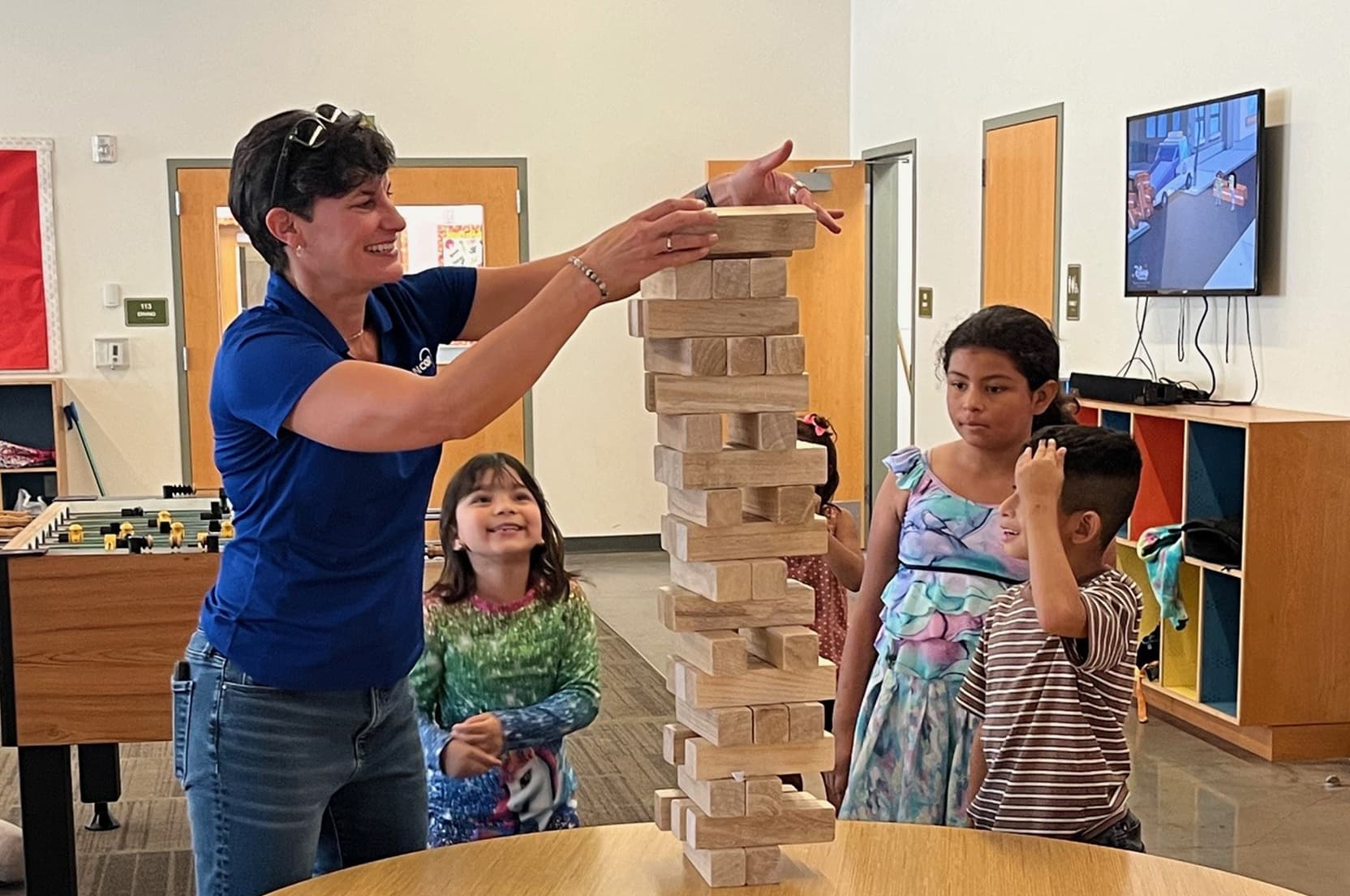 Several children and an adult stand around a round table inside a community room, stacking oversized wooden blocks into a tall tower, with chairs, activity stations and posters visible in the background.