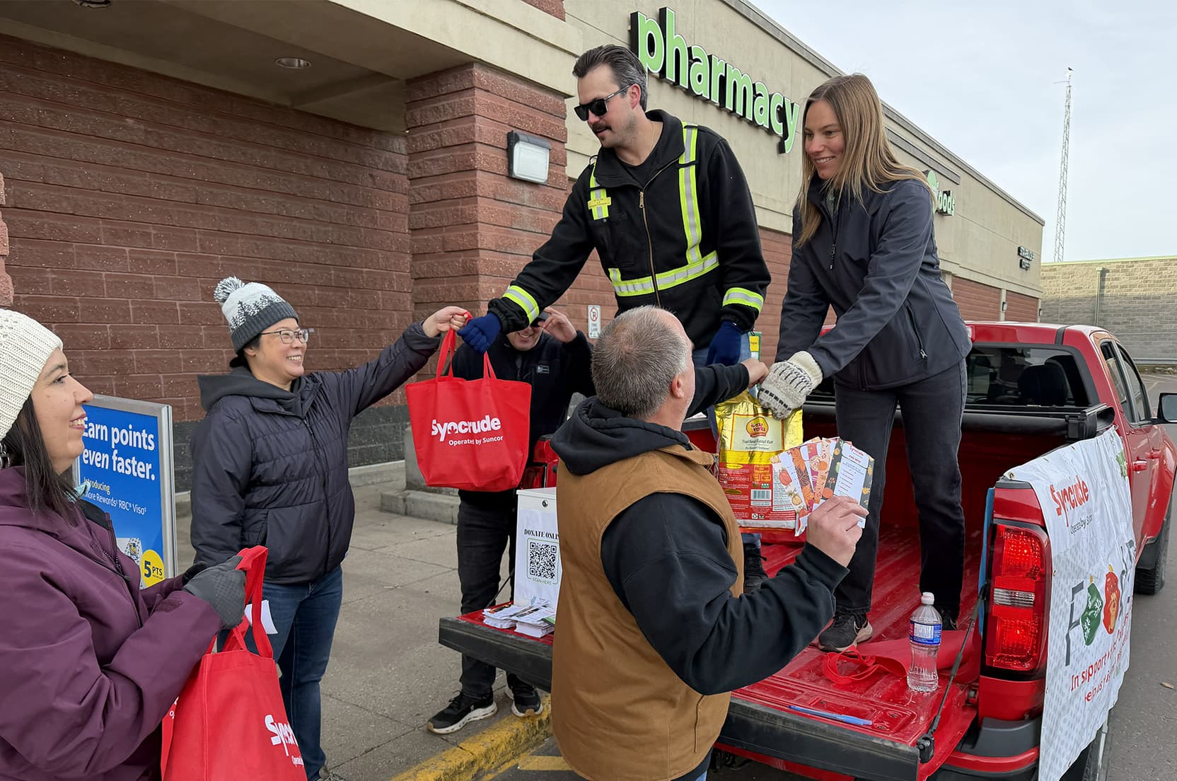 People stand behind a pickup truck outside a building, handing red reusable shopping bags filled with food and supplies from the truck bed to others standing nearby.
