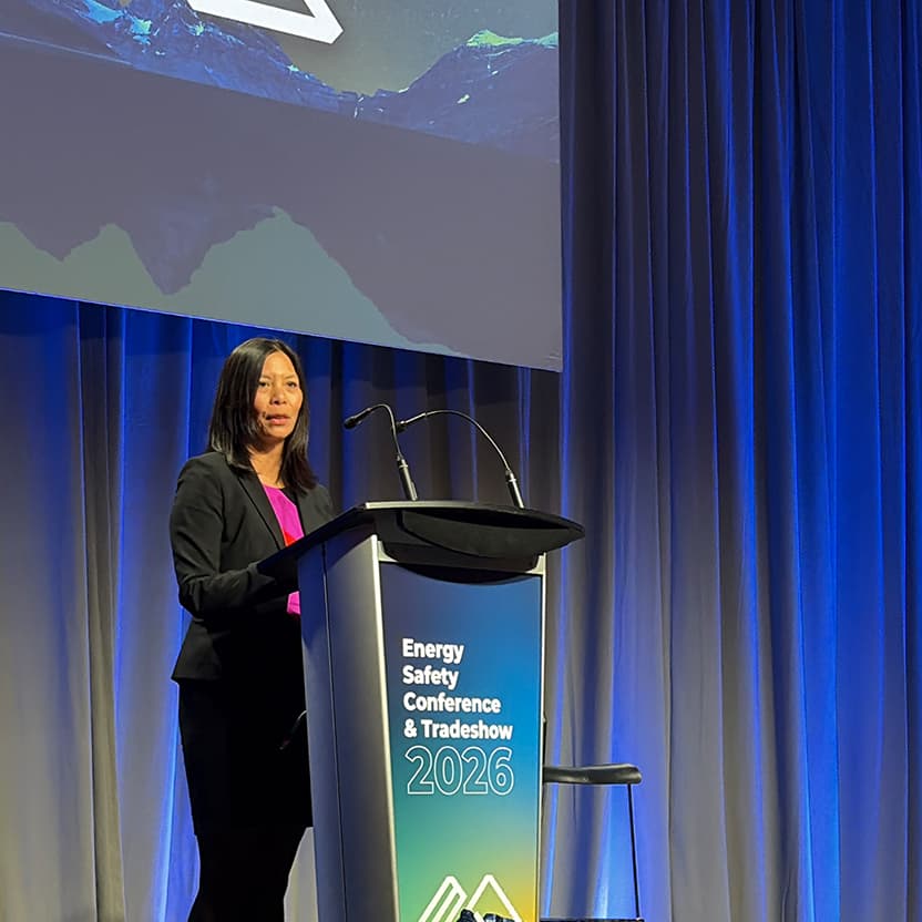 A person stands at a podium on a stage, speaking at the Energy Safety Conference and Tradeshow 2026, with blue stage lighting, curtains and a large presentation screen visible in the background.