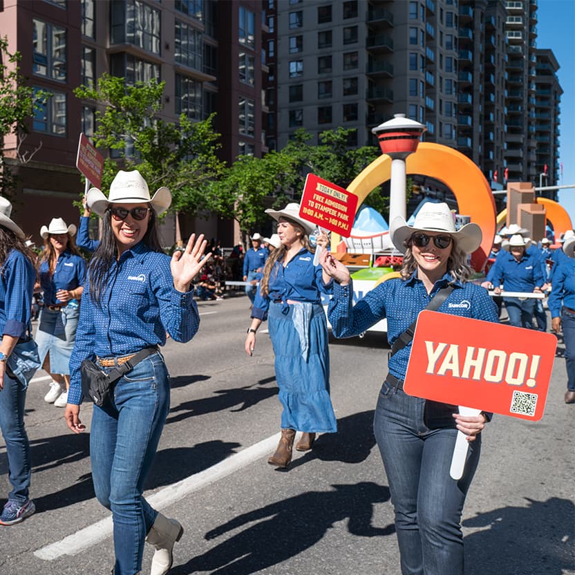 Suncor employees walking in the 2026 Calgary Stampede Parade. They are holding a banner that displays the Calgary Stampede logo beside the Suncor logo, and the Suncor float is behind them.