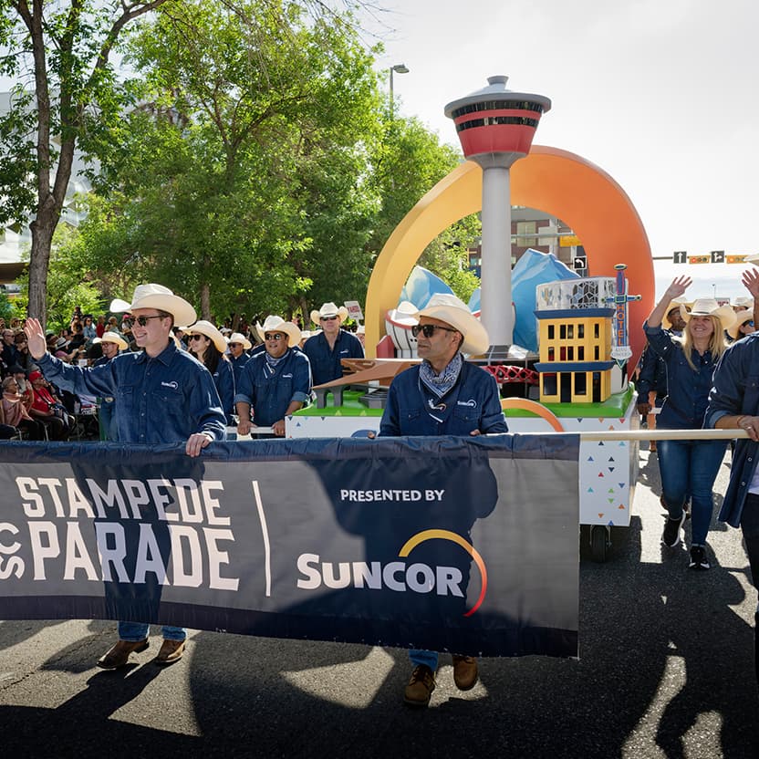 Suncor employees walking in the 2025 Calgary Stampede Parade. They are holding a banner that displays the Calgary Stampede logo beside the Suncor logo, and the Suncor float is behind them.
