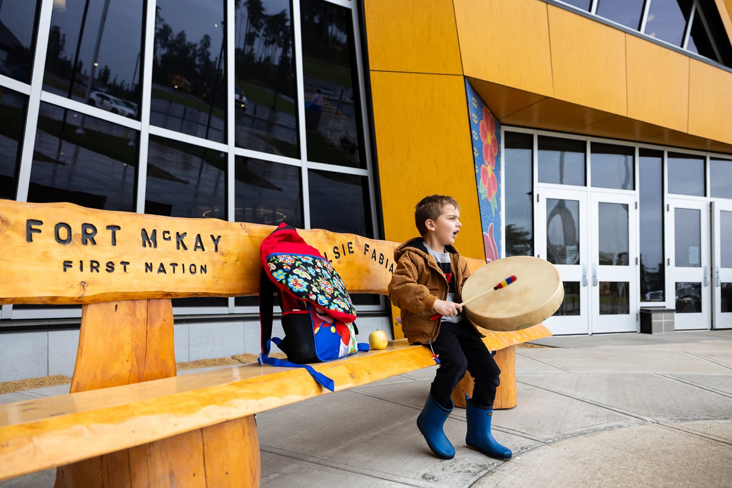 A boy sits on a log-cabin style wooden bench with the inscription “Fort McKay First Nation”, in front of the Elsie Fabian School in Fort McKay, Alberta. He is playing an Indigenous drum and singing.