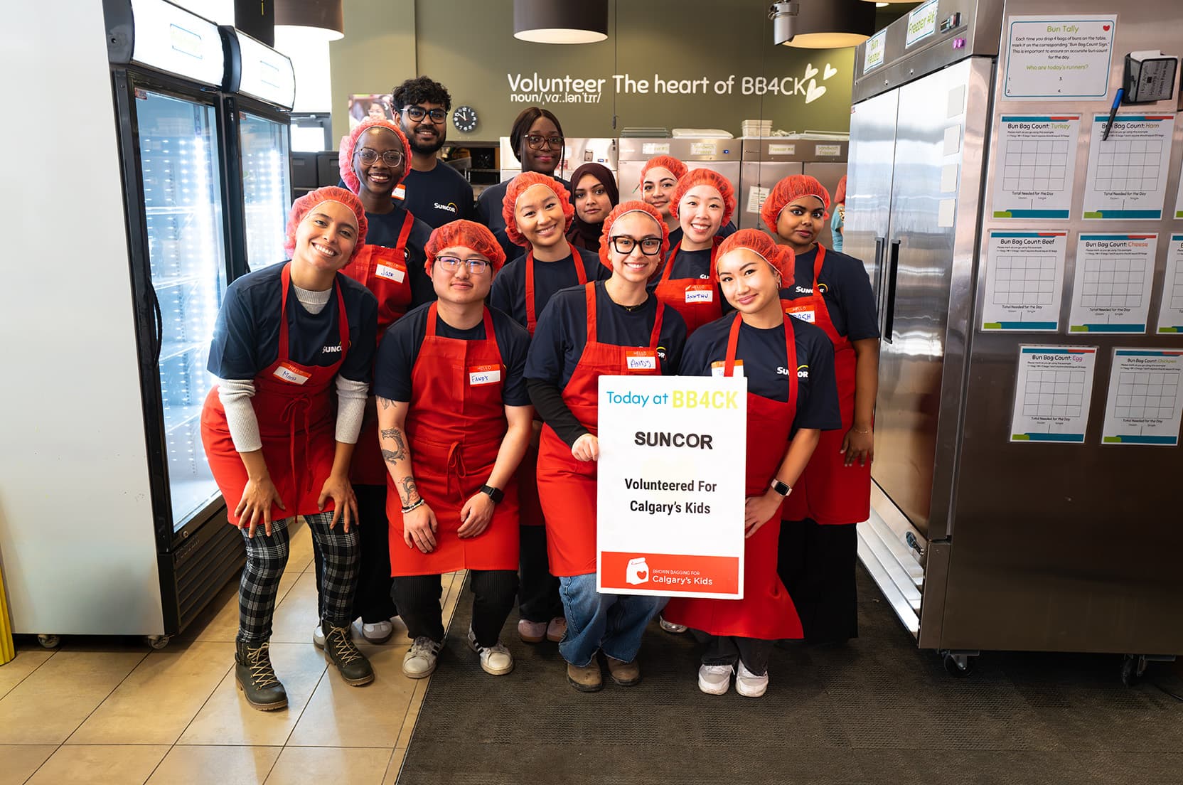 Un groupe de bénévoles portant les mêmes tabliers rouges se tiennent ensemble à l’intérieur d’une zone de distribution ou de dons d’aliments, tenant une grande affiche de remerciement, avec des étagères d’entreposage et des avis affichés, visibles en arrière-plan.