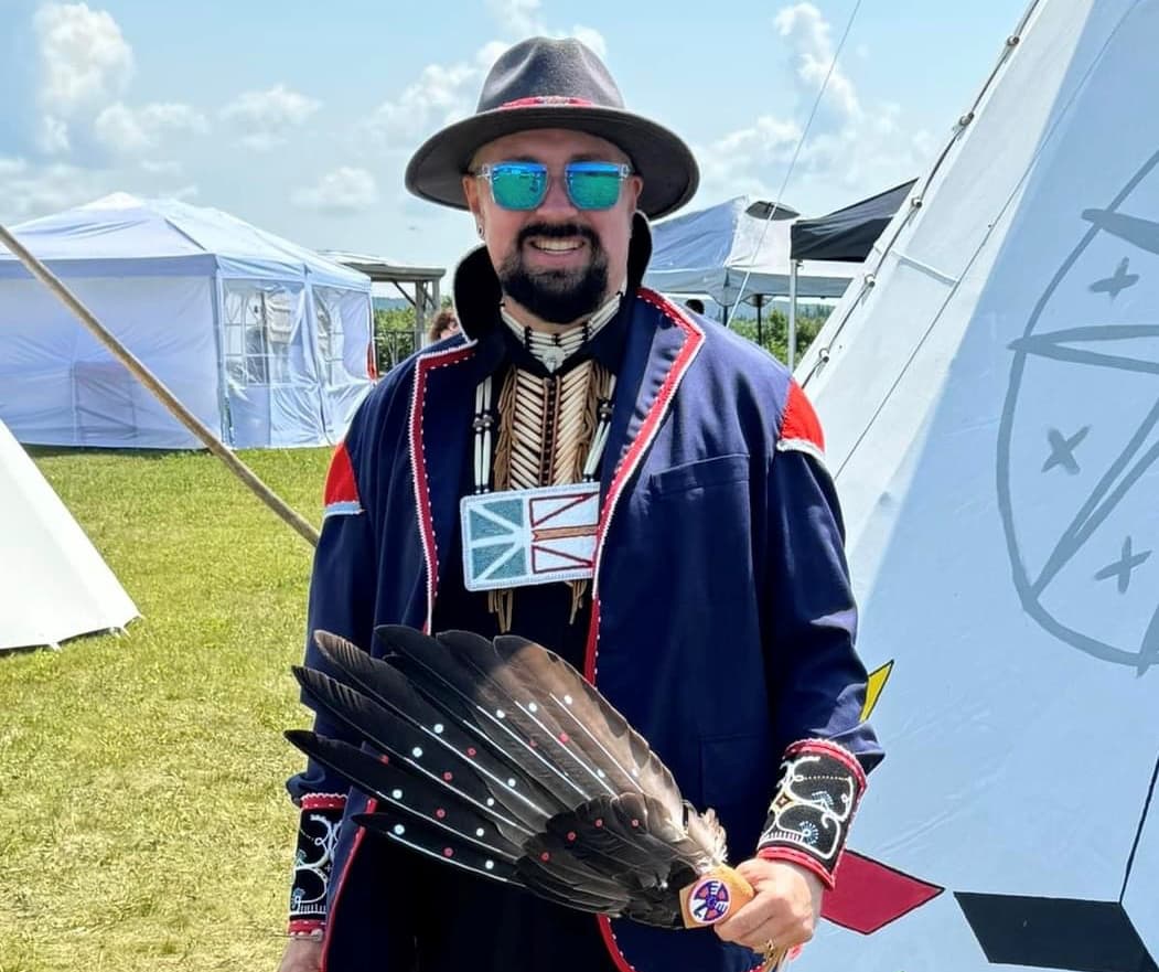 Marcus Gosse sourit, posant pour une photo lors d'un événement extérieur avec des tipis et des chapiteaux en arrière-plan. Il porte un médaillon perlé traditionnel et un chapeau perlé, et tient un éventail en plumes d'aigle sacré.