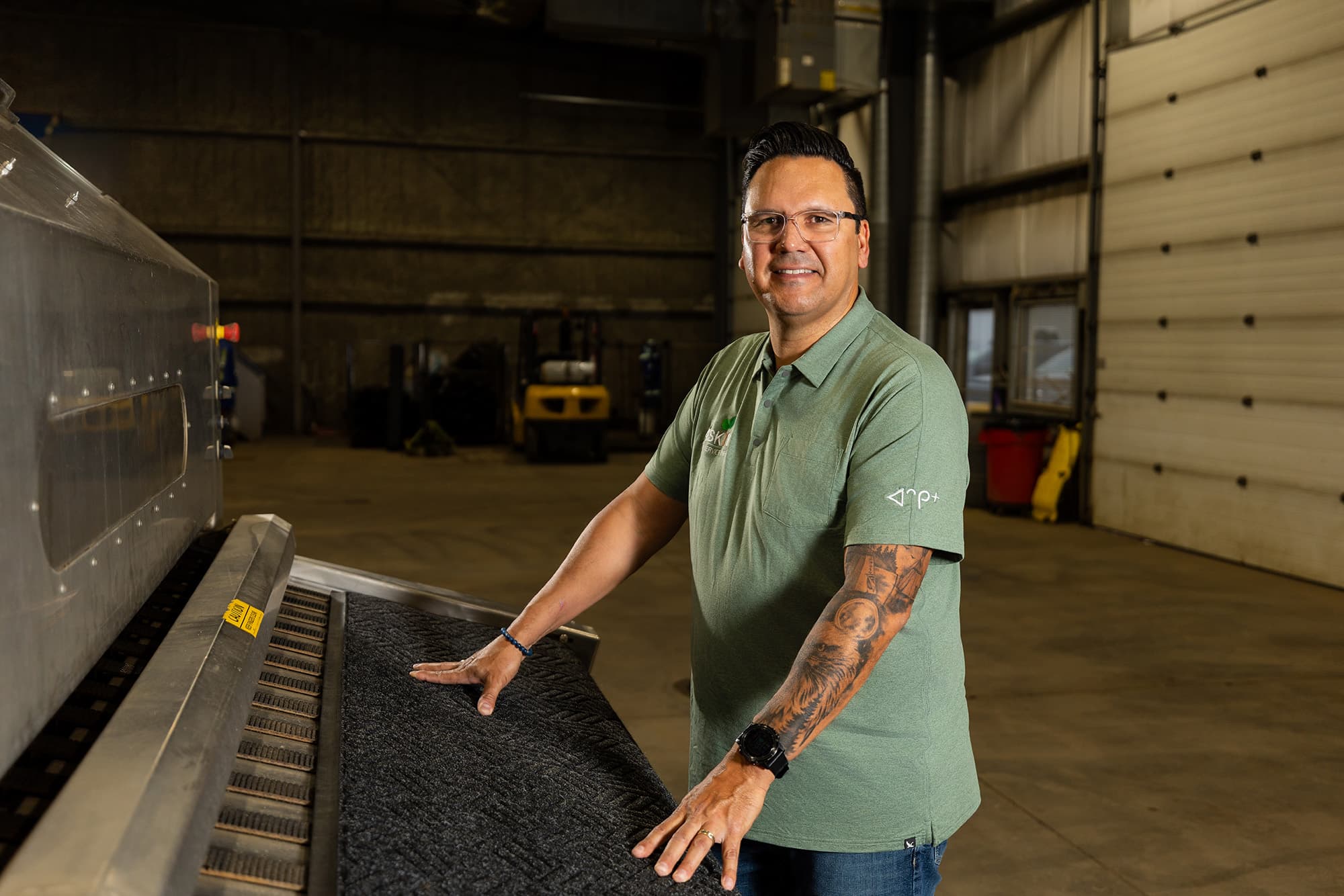 Sandy Sanderson, debout dans l'entrepôt de Askiy Mat Service avec ses mains sur une machine de fabrication de tapis. Il sourit et regarde la caméra.