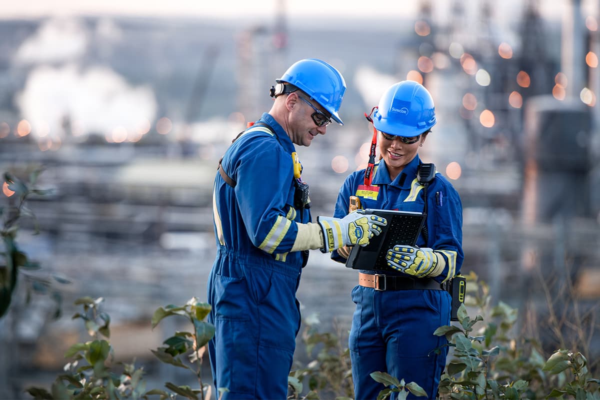 Two workers outside in PPE looking at a tablet together. Base Plant is in the background. 